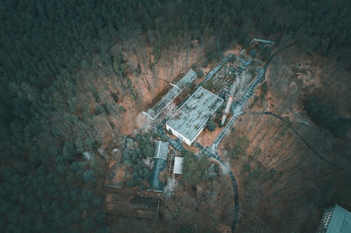 An aerial view of an abandoned industrial complex located within a dense forest. The structures appear to be in a state of decay, with overgrown vegetation surrounding them. The roofs of the buildings are weathered and the paths leading to them are barely visible due to natural overgrowth.