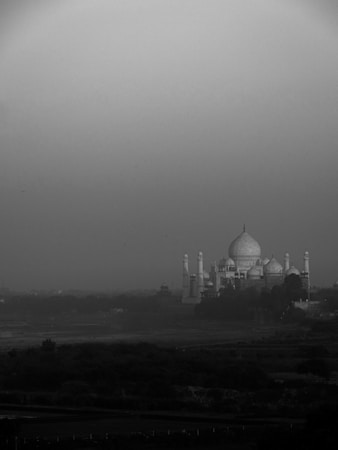 A black and white image depicting the Taj Mahal in the distance, surrounded by a misty or foggy atmosphere. The monument's iconic domes and minarets create a striking silhouette against the muted sky. The foreground features a river or waterway bordered by vegetation, adding depth to the scene.