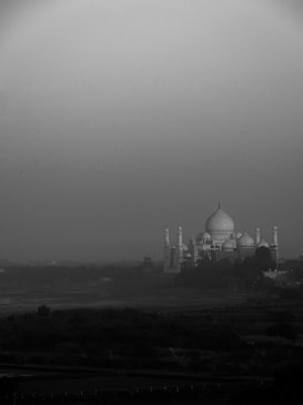 A black and white image depicting the Taj Mahal in the distance, surrounded by a misty or foggy atmosphere. The monument's iconic domes and minarets create a striking silhouette against the muted sky. The foreground features a river or waterway bordered by vegetation, adding depth to the scene.