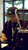 A person wearing a black cap and t-shirt is standing behind a counter, possibly in a café or coffee shop. The individual is interacting with a touchscreen device, with a grinder full of coffee beans nearby. The background features large windows allowing natural light inside.