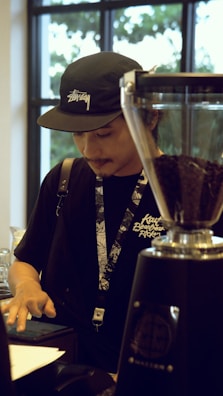 A person wearing a black cap and t-shirt is standing behind a counter, possibly in a café or coffee shop. The individual is interacting with a touchscreen device, with a grinder full of coffee beans nearby. The background features large windows allowing natural light inside.