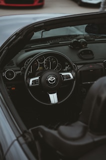 An interior view of a convertible car featuring a sleek steering wheel with a car logo, an instrument cluster with dials, and a manual gear lever. The dashboard has a modern design with climate control buttons and a digital display. There are reflections on the windshield and a glimpse of another car in the background.