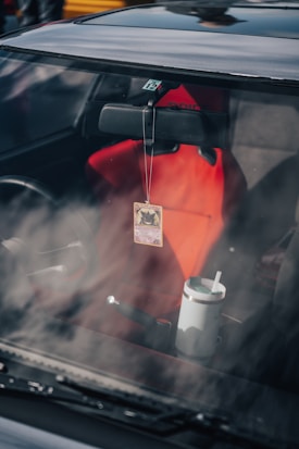 The interior of a car featuring a red seat, a rearview mirror with a card hanging from it, and a large cup with a straw placed near the center console.