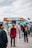 A group of people waiting in line at a colorful coffee truck. The truck has a distinctive orange and turquoise design with various drink images on display. The sky is overcast, and people are dressed in casual, warm clothing, suggesting a cool day.