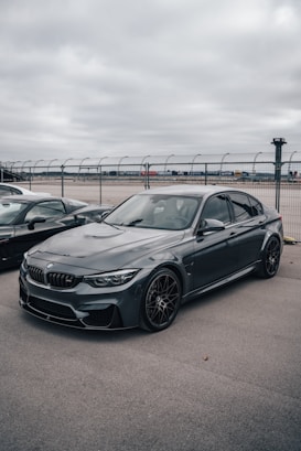 A sleek, dark-colored BMW sedan is parked on a gray tarmac with overcast skies above. The vehicle is positioned in front of a wire fence, suggesting the setting is near an airport or racetrack. The car features sporty rims and a streamlined design.