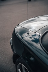 A close-up of the rear section of a dark-colored car displaying a smoothly curved surface. The car's paint reflects a cloudy sky, creating a mirror-like effect. A small antenna is visible on the car's trunk area.