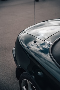 A close-up of the rear section of a dark-colored car displaying a smoothly curved surface. The car's paint reflects a cloudy sky, creating a mirror-like effect. A small antenna is visible on the car's trunk area.