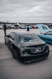 A black car with custom license plate parked in an outdoor car show, surrounded by other vehicles and people. The event is set under a cloudy sky, and the car has tinted windows and stickers on the back.