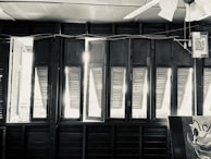 A row of open wooden shutters allowing sunlight to stream into the room. The upper part of a ceiling fan is visible, along with some electrical wires and a hanging picture. The image has a black and white filter, giving it a vintage feel. Part of a painted clown face is in the corner.