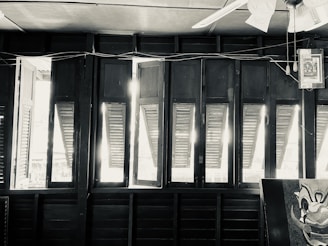 A row of open wooden shutters allowing sunlight to stream into the room. The upper part of a ceiling fan is visible, along with some electrical wires and a hanging picture. The image has a black and white filter, giving it a vintage feel. Part of a painted clown face is in the corner.