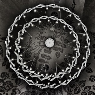 A geometric chandelier with two interlocking circular rings of LED lights is suspended from a ceiling. The intricate ceiling features a dark, floral pattern that adds a dramatic contrast with the bright lights. The chandelier's design is modern and symmetrical, with the lights evenly distributed along the rings.