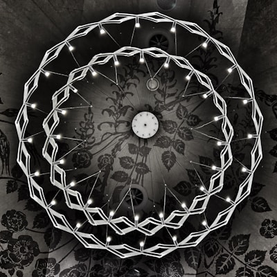 A geometric chandelier with two interlocking circular rings of LED lights is suspended from a ceiling. The intricate ceiling features a dark, floral pattern that adds a dramatic contrast with the bright lights. The chandelier's design is modern and symmetrical, with the lights evenly distributed along the rings.