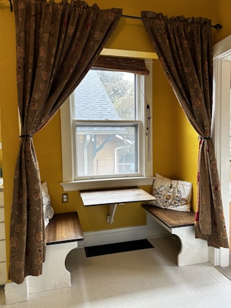 A cozy indoor nook with a small wooden table and benches situated in front of a window. The space is decorated with patterned curtains on either side of the window and a few decorative cushions on the benches. The walls are painted in a warm yellow shade, and natural light is streaming through the window.