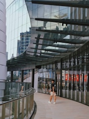 A modern urban setting with a curved glass facade reflecting surrounding buildings. A person wearing shorts and a backpack walks along a tiled pathway adjacent to the glass wall. The architecture features a combination of metal and glass materials, showcasing a contemporary design.