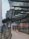 A modern urban setting with a curved glass facade reflecting surrounding buildings. A person wearing shorts and a backpack walks along a tiled pathway adjacent to the glass wall. The architecture features a combination of metal and glass materials, showcasing a contemporary design.