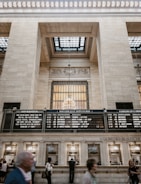 A bustling train station hall features high ceilings with ornate architectural details and a large window grid allowing natural light to filter through. Hanging from the ceiling is an elegant chandelier. Below, a busy information and ticket counter is attended by people. LED departure boards indicate train schedules for the Harlem Line.