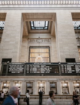 A bustling train station hall features high ceilings with ornate architectural details and a large window grid allowing natural light to filter through. Hanging from the ceiling is an elegant chandelier. Below, a busy information and ticket counter is attended by people. LED departure boards indicate train schedules for the Harlem Line.