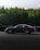 A sleek black sports car is in motion on a race track, surrounded by a green forest. The car's number 911 is visible on the side window, highlighting its participation in a racing event. The road surface is dark, and the overall scene conveys a sense of speed and focus.