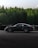 A sleek black sports car is in motion on a race track, surrounded by a green forest. The car's number 911 is visible on the side window, highlighting its participation in a racing event. The road surface is dark, and the overall scene conveys a sense of speed and focus.
