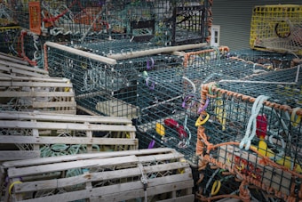 A collection of lobster traps made from metal and wooden materials is stacked outdoors. The traps are intricately connected with ropes of various colors and are equipped with colorful flotation devices used for identification. Some traps are constructed from wire mesh with a rusty appearance, and others are wooden with netting stretched across them. The scene suggests a coastal or fishing environment.
