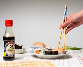 A hand is using chopsticks to pick up a piece of salmon nigiri from a plate of sushi, which includes various sushi rolls. A bottle of soy sauce with a red cap is placed to the left of the plate. The table is set with a woven mat and a patterned cloth, creating a neat and inviting dining setup.