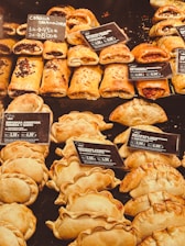 An assortment of baked pastries and empanadas on display with labels indicating different types and prices. The pastries are neatly arranged in rows, showcasing various fillings and crusts. The warm lighting emphasizes the golden-brown color of the baked goods.