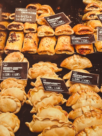 An assortment of baked pastries and empanadas on display with labels indicating different types and prices. The pastries are neatly arranged in rows, showcasing various fillings and crusts. The warm lighting emphasizes the golden-brown color of the baked goods.
