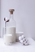 A still life composition featuring a glass bottle filled with milk, closed with a cork stopper, placed next to a double-walled glass containing more milk. Cotton flowers are elegantly arranged around the bottle and glass, creating a soft and natural atmosphere. A white bowl sits in the background, adding to the minimalist aesthetic.