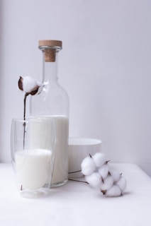 A still life composition featuring a glass bottle filled with milk, closed with a cork stopper, placed next to a double-walled glass containing more milk. Cotton flowers are elegantly arranged around the bottle and glass, creating a soft and natural atmosphere. A white bowl sits in the background, adding to the minimalist aesthetic.