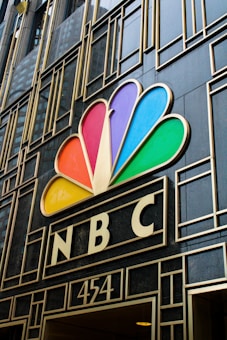 A building facade featuring the colorful peacock logo of NBC prominently displayed above the entrance. The design includes geometric patterns and the number 454 below the logo.