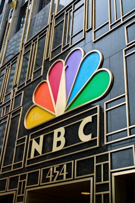 A building facade featuring the colorful peacock logo of NBC prominently displayed above the entrance. The design includes geometric patterns and the number 454 below the logo.