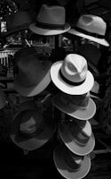 Multiple hats are arranged on a stand or display rack, with a variety of styles and textures. The scene is outdoors and shaded, and sunlight creates contrasts and shadows on the different hats.