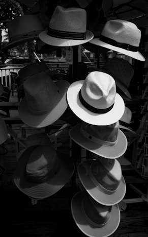 Multiple hats are arranged on a stand or display rack, with a variety of styles and textures. The scene is outdoors and shaded, and sunlight creates contrasts and shadows on the different hats.