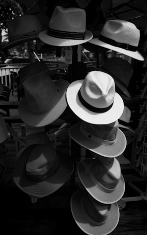 Multiple hats are arranged on a stand or display rack, with a variety of styles and textures. The scene is outdoors and shaded, and sunlight creates contrasts and shadows on the different hats.