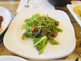 A fresh salad composed of various leafy greens, carrot slices, and topped with sesame seeds. The salad is neatly arranged on a white plate. Adjacent dishes and a wooden table are partially visible.