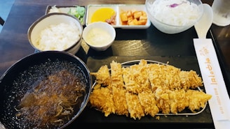 A meal featuring breaded and fried cutlet served on a rectangular tray, accompanied by a bowl of soup with noodles, white rice in a separate bowl, and various side dishes including cabbage salad, yellow pickles, cubed kimchi, and wasabi. There is also a pair of chopsticks with Korean text.