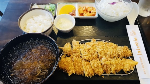 A meal featuring breaded and fried cutlet served on a rectangular tray, accompanied by a bowl of soup with noodles, white rice in a separate bowl, and various side dishes including cabbage salad, yellow pickles, cubed kimchi, and wasabi. There is also a pair of chopsticks with Korean text.