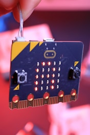 A microcontroller board with an array of LED lights lit up in a pattern. The board has two buttons labeled A and B, and features connectors at the bottom. The background is blurred, with red and pink tones.
