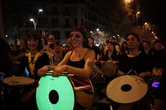 A group of people is participating in a nighttime event, playing various drums and percussion instruments. They are dressed in dark clothing with some face paint and tattoos. The background reveals a street setting with buildings and street lights.