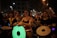 A group of people is participating in a nighttime event, playing various drums and percussion instruments. They are dressed in dark clothing with some face paint and tattoos. The background reveals a street setting with buildings and street lights.