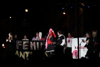 A group of people is gathered on a stage at night, holding various signs and banners. One person in the center holds a microphone and wears a red top and pink scarf. Some individuals are gesturing as if in sign language, while others hold banners with colorful lettering and images.