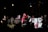 A group of people is gathered on a stage at night, holding various signs and banners. One person in the center holds a microphone and wears a red top and pink scarf. Some individuals are gesturing as if in sign language, while others hold banners with colorful lettering and images.