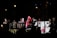 A group of people is gathered on a stage at night, holding various signs and banners. One person in the center holds a microphone and wears a red top and pink scarf. Some individuals are gesturing as if in sign language, while others hold banners with colorful lettering and images.