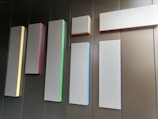 Several rectangular panels of varying lengths and widths are mounted on a wall. Each panel has a slight glow on one of its edges, with different colors such as yellow, red, green, blue, and orange. The wall behind the panels appears to be made of dark brown or black material, creating a minimalist and modern aesthetic.