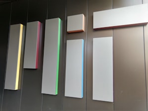 Several rectangular panels of varying lengths and widths are mounted on a wall. Each panel has a slight glow on one of its edges, with different colors such as yellow, red, green, blue, and orange. The wall behind the panels appears to be made of dark brown or black material, creating a minimalist and modern aesthetic.