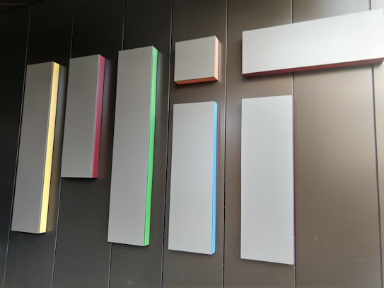 Several rectangular panels of varying lengths and widths are mounted on a wall. Each panel has a slight glow on one of its edges, with different colors such as yellow, red, green, blue, and orange. The wall behind the panels appears to be made of dark brown or black material, creating a minimalist and modern aesthetic.