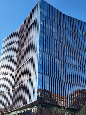 A modern glass office building with reflective windows and sleek architectural lines. The structure features a grid-like pattern of vertical and horizontal elements. The sky is clear and blue, enhancing the building's reflective qualities.
