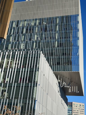 A modern multi-story building with a facade made up of vertical glass and metal panels. The structure features a unique overhang and glass windows providing a reflective surface. The sign on the facade suggests it is a commercial or office building. In the background, a clear blue sky can be seen.
