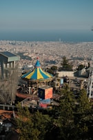 An amusement park with a colorful carousel in the foreground, surrounded by trees and visitors. Beyond the park, a sprawling cityscape stretches out to meet the horizon, with the sea in the distance and clear blue skies overhead.