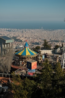 An amusement park with a colorful carousel in the foreground, surrounded by trees and visitors. Beyond the park, a sprawling cityscape stretches out to meet the horizon, with the sea in the distance and clear blue skies overhead.